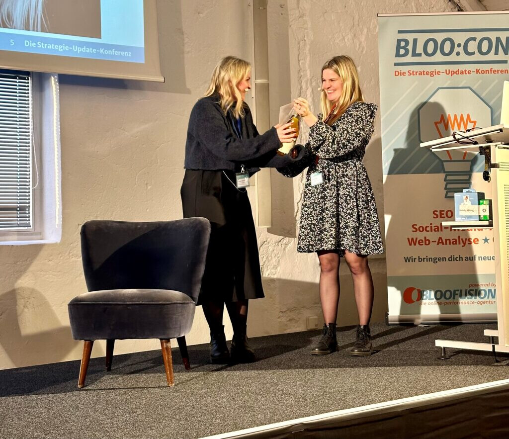 Zwei Frauen stehen auf der Bühne der BLOO:CON-Konferenz 2024. Eine Frau überreicht der anderen ein Geschenk. Beide lächeln. Im Hintergrund ist ein BLOO:CON-Banner mit dem Schriftzug zu sehen.