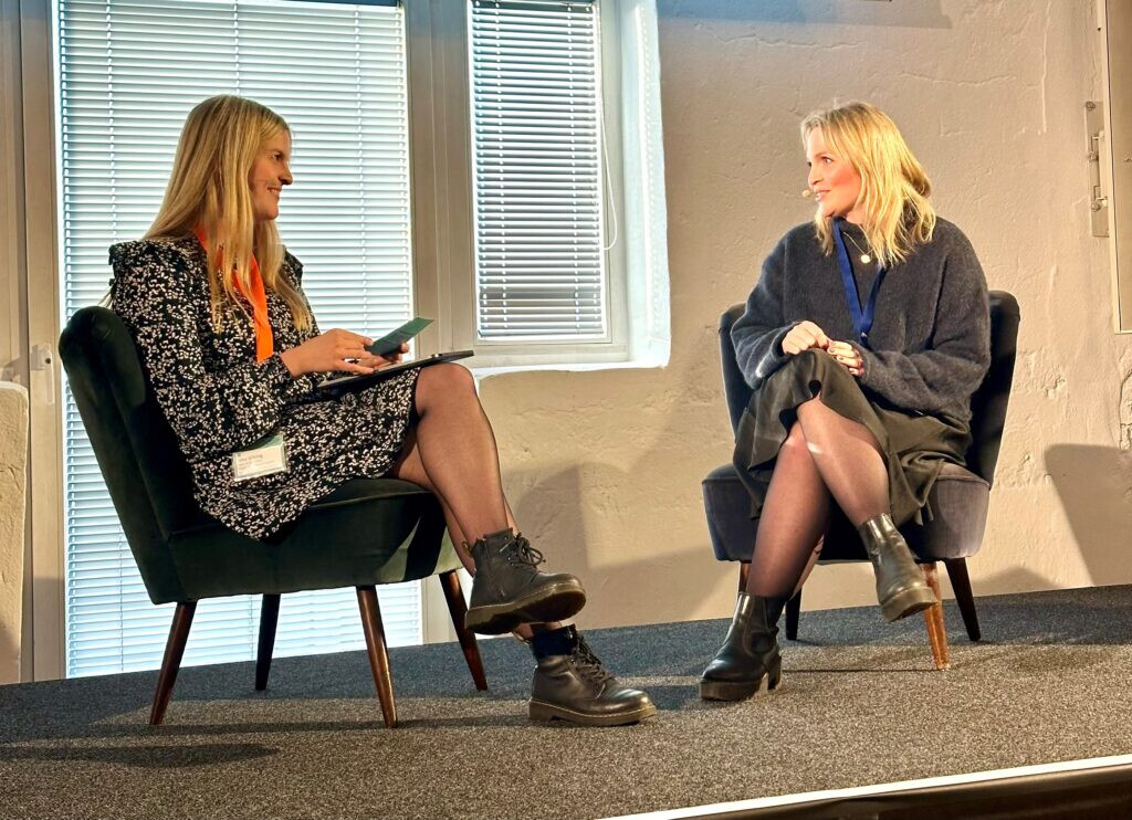 Zwei Frauen sitzen auf einer Bühne in Sesseln und führen ein Interview. Beide lächeln sich an. Im Hintergrund fällt Tageslicht durch ein Fenster. Aufgenommen bei der BLOO:CON-Konferenz 2024.