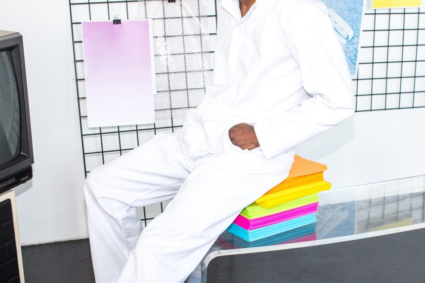 Eine Person in weißer Kleidung sitzt auf einem transparenten Tisch, unter dem bunte Papierstapel in Neonfarben liegen. Im Hintergrund sind Gitterwände mit aufgehängten Farbmusterblättern und ein alter Fernseher zu sehen. Die Szene wirkt modern und minimalistisch.