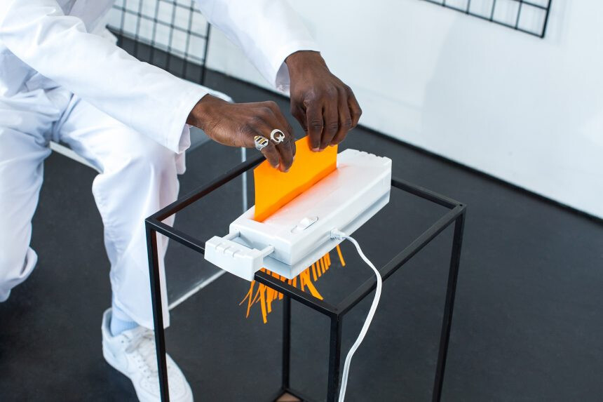 Eine Person in weißer Kleidung steckt ein leuchtend orangefarbenes Blatt Papier in einen weißen Aktenvernichter auf einem schwarzen Metalltisch. Das Papier wird gerade zerkleinert, orangefarbene Streifen fallen heraus. Der Raum ist minimalistisch eingerichtet mit weißen Wänden und dunklem Boden.