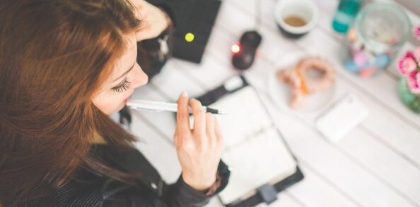 Eine Frau sitzt an einem Schreibtisch, hält nachdenklich einen Stift an die Lippen und blickt in ein aufgeschlagenes Notizbuch. Auf dem Tisch liegen Laptop, Maus, Kaffeetasse, Gebäck und Blumen.