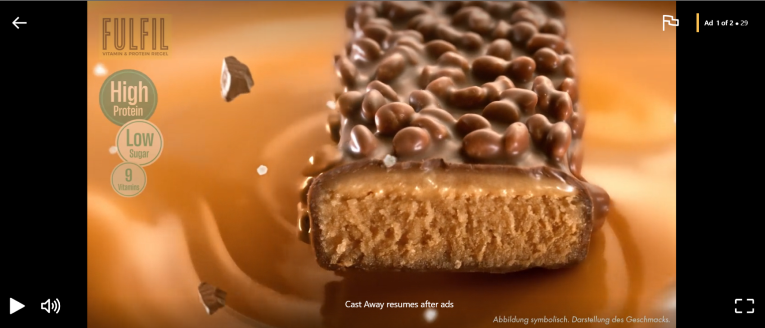 Nahaufnahme eines Fulfil Vitamin- & Proteinriegels, der in flüssigem Karamell liegt. Der Riegel ist zur Hälfte angeschnitten und zeigt eine cremige Füllung unter einer Schicht aus Schokolade und knusprigen Stückchen. Links stehen die Hinweise „High Protein“, „Low Sugar“ und „9 Vitamins“.